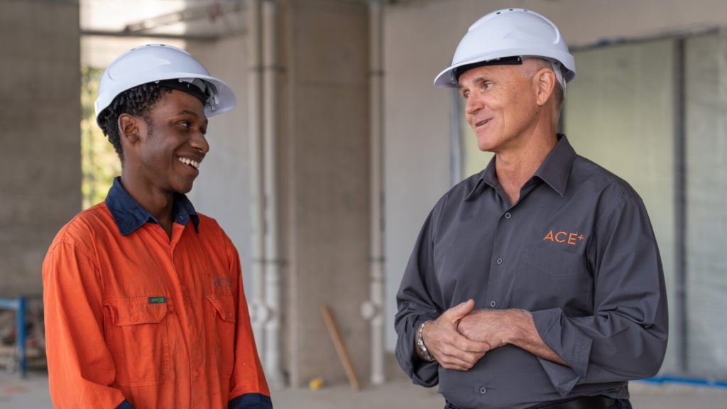 Two men wearing white hard hats and talking to one another whilst smiling 