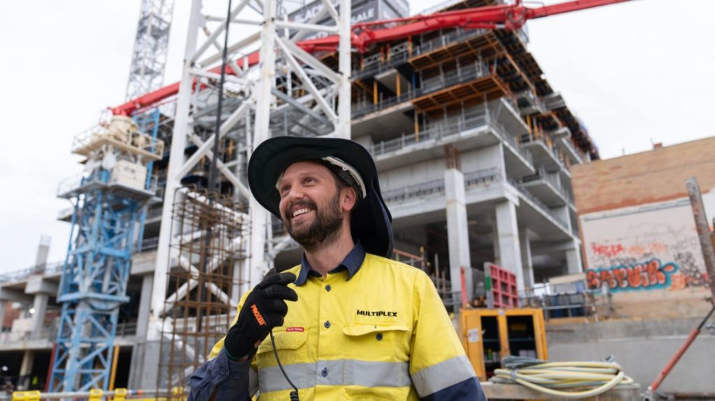 Man in yellow trade uniform holding radio receiver and standing in front of building in progress