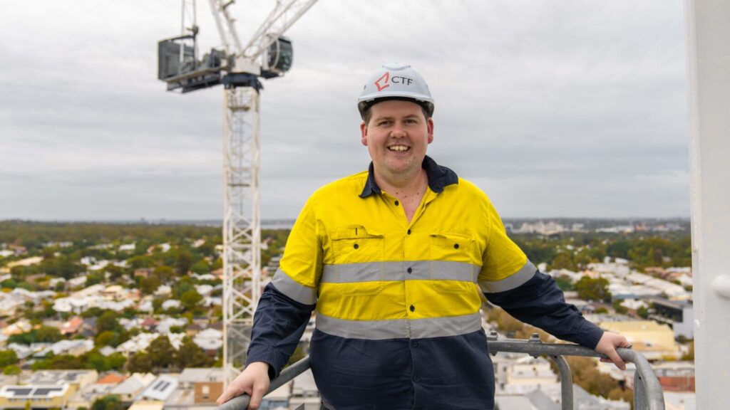 Construction workers wearing a hardhat and standing at the top of a building with a crane in the background