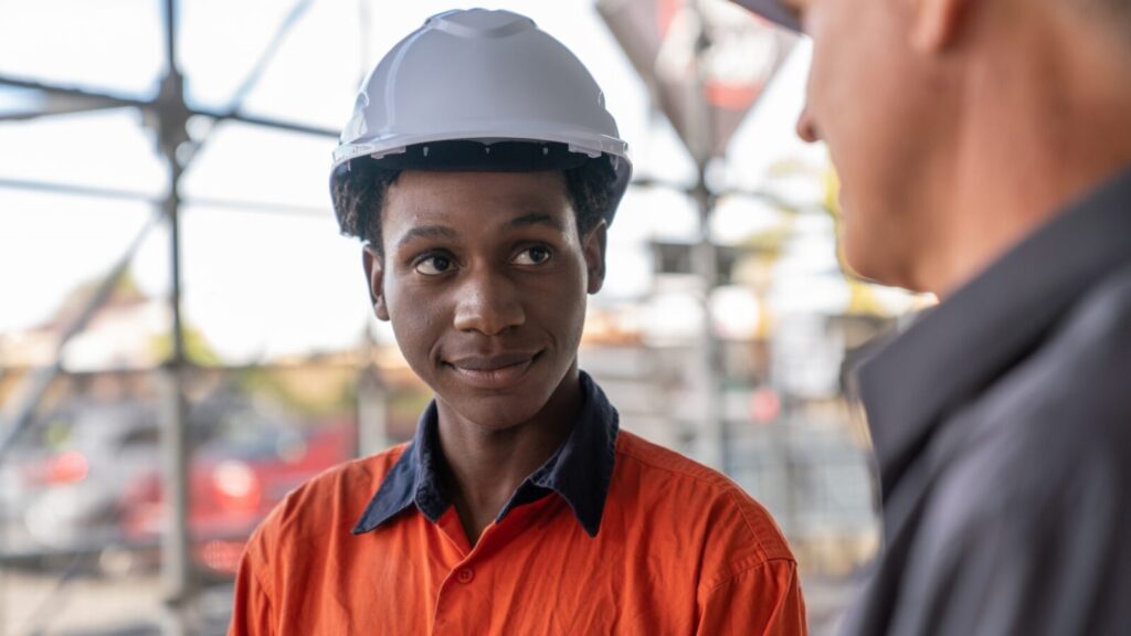 Apprentice in high vis and white hard hat listening to man in grey shirt