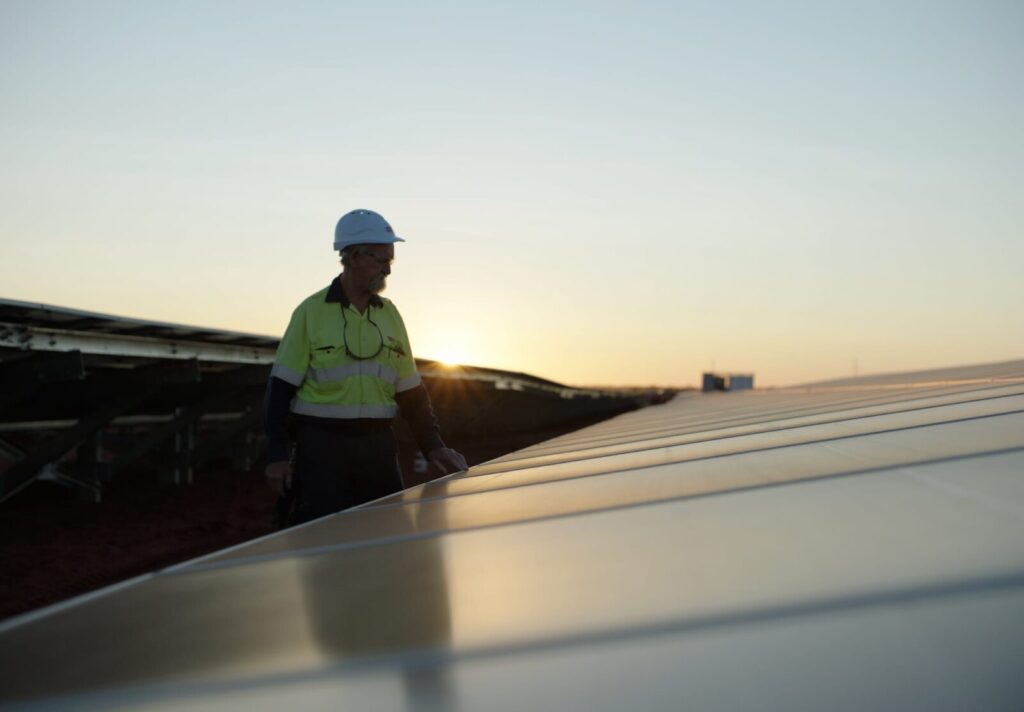Worker in a hard hat and safety gear inspects solar panels at sunset, with a clear sky and horizon in the background.
