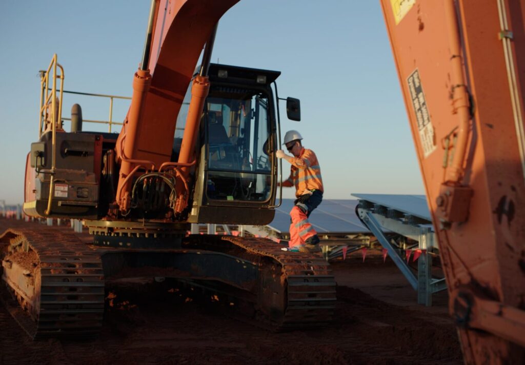 A construction worker in a hard hat and safety gear stands on an excavator at a construction site with solar panels in the background.