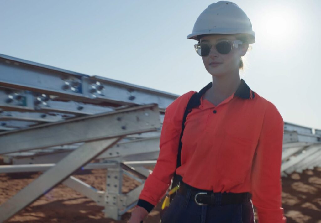 Person in a bright orange safety shirt and hard hat, wearing sunglasses, walking near metal structures at a construction site. Sunlight in the background.